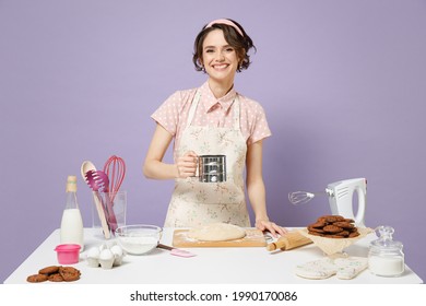 Young Fun Housewife Housekeeper Chef Baker Woman In Pink Apron Work At Table Kitchenware Use Flour Sifter Mug Baking Cake Look Camera Isolated On Pastel Violet Background Process Cooking Food Concept.