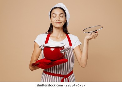 Young fun housewife housekeeper chef baker latin woman wear striped apron toque hat hold in hand red pot pan sniff aroma dish meal isolated on plain pastel light beige background. Cook food concept - Powered by Shutterstock