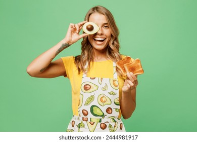 Young fun housewife housekeeper chef cook baker woman wear apron yellow t-shirt cover eye with half of avocado hold in hand bread toast isolated on plain green background studio. Cooking food concept - Powered by Shutterstock