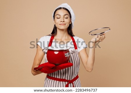 Young fun housewife housekeeper chef baker latin woman wear striped apron toque hat hold in hand red pot pan sniff aroma dish meal isolated on plain pastel light beige background. Cook food concept