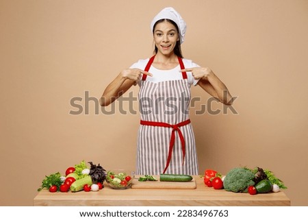 Young fun housewife housekeeper chef cook latin woman in apron toque chefs hat work at table kitchenware point fingers on herself isolated on plain pastel light beige background. Process cooking food