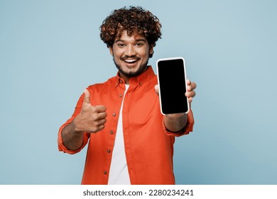 Young fun Indian man wears orange red shirt white t-shirt hold in hand use close up mobile cell phone with blank screen workspace area show thumb up isolated on plain pastel light blue cyan background