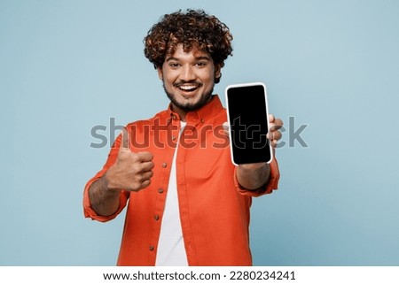 Young fun Indian man wears orange red shirt white t-shirt hold in hand use close up mobile cell phone with blank screen workspace area show thumb up isolated on plain pastel light blue cyan background