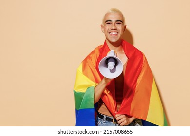 Young fun latin gay man with make up in beige tank shirt wrapped in rainbow flag hold shout aside in megaphone isolated on plain light ocher background studio portrait People lgbt lifestyle concept