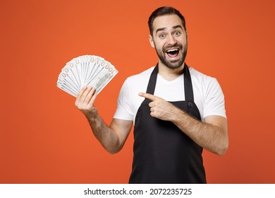 Young Fun Man 20s Barista Bartender Barman Employee Wearing Black Apron White T-shirt Work In Coffee Shop Point Finger On Fan Of Money In Dollars Isolated On Orange Background. Small Business Startup.