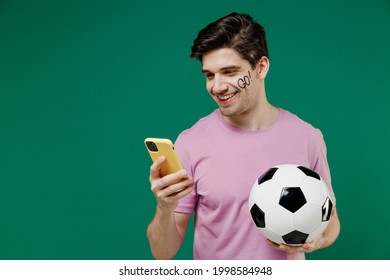 Young fun man 20s fan wear basic pink t-shirt cheer up support football sport team hold in hand mobile cell phone soccer ball watch tv live stream scream isolated on dark green color background studio - Shutterstock ID 1998584948
