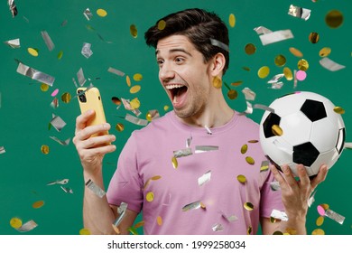 Young fun man 20s fan wear basic pink t-shirt cheer up support football sport team hold in hand mobile cell phone soccer ball watch tv live stream scream isolated on dark green color background studio - Shutterstock ID 1999258034