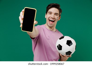 Young fun man 20s fan wear basic pink t-shirt cheer up support football sport team hold in hand mobile cell phone soccer ball watch tv live stream scream isolated on dark green color background studio