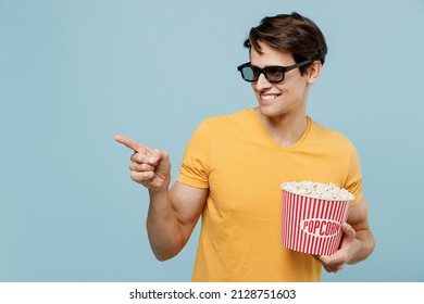Young Fun Man In 3d Glasses Watch Movie Film Hold Bucket Of Popcorn Point Index Finger Aside On Area Isolated On Plain Pastel Light Blue Background Studio. People Emotions In Cinema Lifestyle Concept