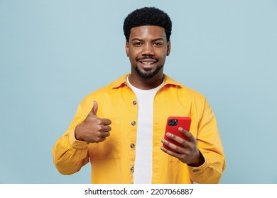 Young Fun Man Of African American Ethnicity 20s In Yellow Shirt Hold In Hand Use Mobile Cell Phone Show Thumb Up Isolated On Plain Pastel Light Blue Background Studio Portrait People Lifestyle Concept