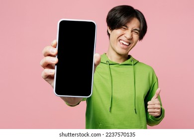 Young fun man of Asian ethnicity wear green hoody look camera hold in hand use close up mobile cell phone with blank screen workspace area show thumb up isolated on plain pastel light pink background - Powered by Shutterstock