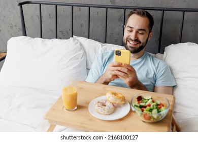 Young Fun Man In Blue T-shirt He Lying In Bed Have Breakfast Taking Picture Shot On Mobile Cell Phone Of Food Rest Relax Spend Time In Bedroom Lounge Home In Own Room House. Good Mood Bedtime Concept