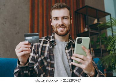 Young Fun Man He Wears Brown Shirt Using Mobile Cell Phone Hold Credit Bank Card Shopping Online Order Delivery Booking Tour Sitting On Blue Sofa In Own Living Room Apartment Stay Home Indoor Flat On