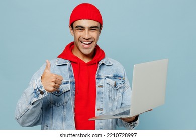 Young fun middle eastern IT man 20s he wear denim jacket red hat hold use work on laptop pc computer show thumb up isolated on plain pastel light blue cyan background studio. People lifestyle concept