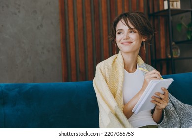 Young Fun Minded Pensive Woman Wrapped In Plaid Write Down Memories In Notebook Diary Sit On Blue Sofa Stay At Home Flat Rest Relax Spend Free Spare Time In Living Room Indoors People Lounge Concept