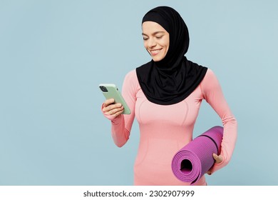 Young fun muslim fitness trainer sporty woman wears pink abaya hijab spend time in home gym hold yoga mat use mobile cell phone isolated on plain blue background studio Workout sport fit abs concept - Powered by Shutterstock