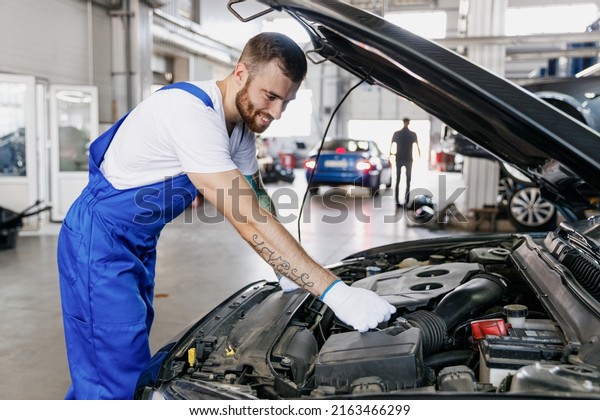 Young fun professional technician car mechanic man in\
denim blue overalls white t-shirt gloves fixing problem with raised\
hood work in modern vehicle repair shop workshop indoor Tattoo\
translate fun