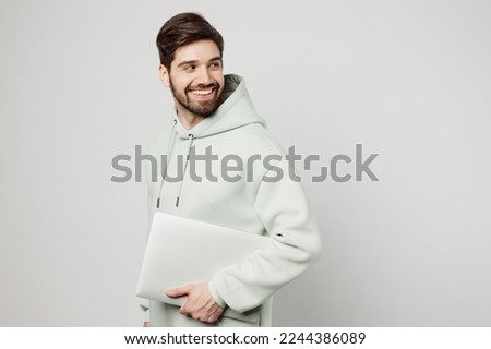 Young fun programmer happy caucasian man wear mint hoody hold closed laptop pc computer look aside on workspace area isolated on plain solid white background studio portrait. People lifestyle concept