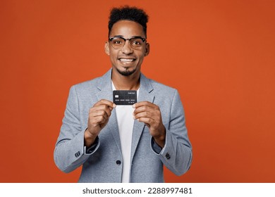 Young fun rich employee business man corporate lawyer wear classic formal grey suit shirt glasses work in office hold in hand mock up of credit bank card isolated on plain red orange background studio