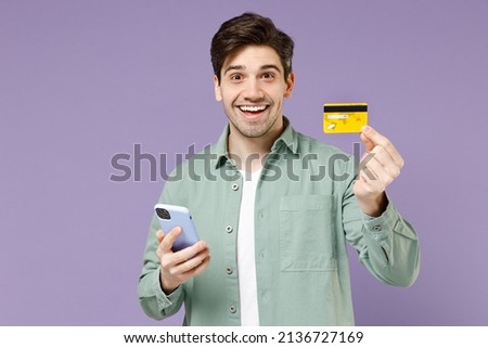 Young fun satisfied caucasian man 20s in casual mint shirt white t-shirt hold mobile cell phone credit bank card shopping online isolated on purple background studio portrait People lifestyle concept