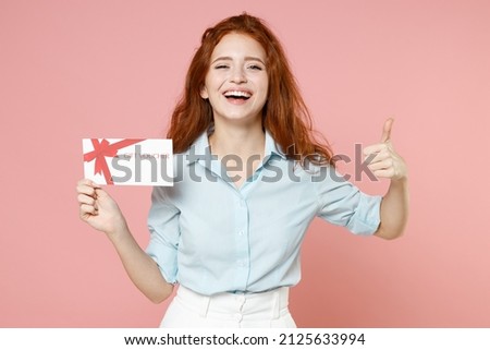 Young fun satisfied happy ginger student redhead woman 20s wearing blue shirt holding gift voucher flyer mock up showing thumb up like gesture isolated on pastel pink color background studio portrait
