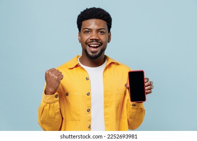 Young fun satisfied man of African American ethnicity 20s wear yellow shirt hold use mobile cell phone with blank screen workspace area do winner gesture isolated on plain pastel light blue background - Shutterstock ID 2252693981