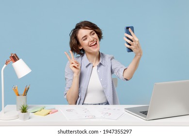 Young fun secretary employee business woman in casual shirt sit work at white office desk with pc laptop do selfie shot on mobile phone show victory v-sign gesture isolated on blue background studio