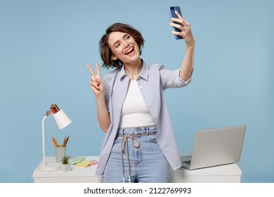Young fun secretary employee business woman in casual shirt work stand at white office desk with pc laptop do selfie shot on mobile phone show victory v-sign gesture isolated on blue background studio