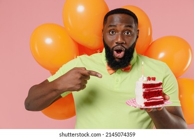 Young fun shocked black man 20s in green t-shirt bow tie hold bunch of air inflated helium balloons celebrate birthday party point finger on cake with candle isolated on plain pastel pink background.