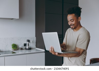 Young Fun Smiling Confident Freelancer African American Man 20s In Casual Clothes Hold Use Work On Laptop Pc Computer Browsing Internet In Light Kitchen At Home Alone Indoor. People Lifestyle Concept.