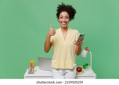 Young Fun Smiling Employee Business Woman Of African American Ethnicity In Shirt Stand Work At White Office Desk With Pc Laptop Hold Mobile Cell Phone Show Thumb Up Isolated On Plain Green Background.