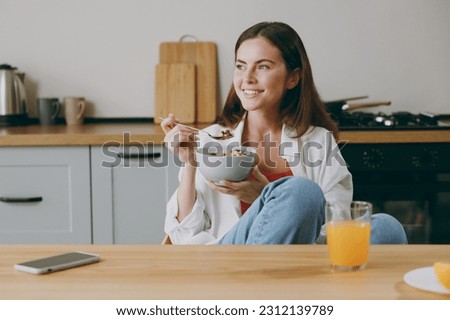 Young fun smiling european happy housewife woman wear casual clothes look aside eat breakfast muesli cereals with milk fruit in bowl cooking food in light kitchen at home alone. Healthy diet concept