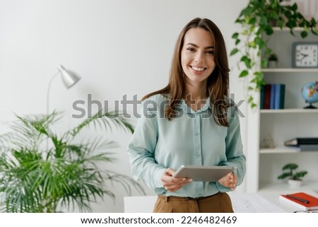 Young fun smiling european successful employee business woman in blue shirt work use hold tablet pc computer stand at workplace white desk at light modern office indoors. Achievement career concept