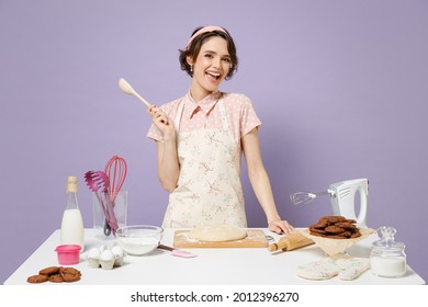 Junge lustige Lächeln glückliche Hausfrau Hausfrau Haushälterin Koch Chef Bäcker Frau in rosafarbenen Vorhängen am Tisch Küchengeschirr halten Holzlöffel einzeln auf pastellviolettem Hintergrund Studioprozess Kochkonzept