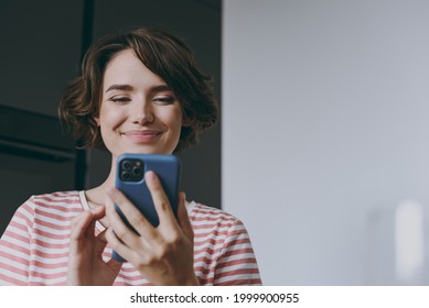 Young fun smiling housewife woman 20s wearing casual clothes striped t-shirt use mobile cell phone chatting online browsing surfing internet in light kitchen at home alone People lifestyle concept.