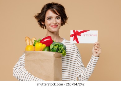 Young Fun Smiling Vegetarian Woman 20s In Casual Clothes Hold Paper Bag With Vegetables Hold Gift Certificate Coupon Voucher Card For Store Isolated On Plain Pastel Beige Background Shopping Concept