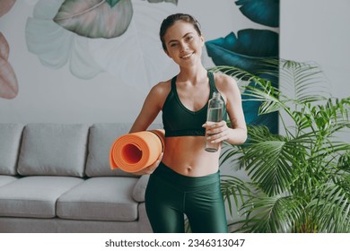 Young fun strong sporty athletic fitness trainer instructor woman wearing green tracksuit hold in hand yoga mat drink water training do exercises at home gym indoor. Workout sport motivation concept