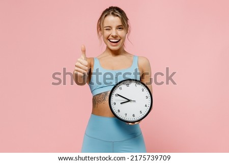 Young fun strong sporty athletic fitness trainer instructor woman wear blue tracksuit spend time in home gym hold clock show thumb up isolated on pastel plain pink background. Workout sport concept