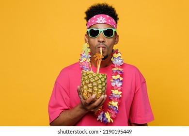 Young fun stylish man 20s he wearing pink t-shirt sunglasses hawaiian lei near hotel pool drink straw pineapple juice look camera isolated on plain yellow background. Summer vacation sea rest concept