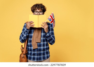 Young fun teen Indian boy IT student he wear casual clothes shirt glasses bag cover mouth with book hold British flag isolated on plain yellow color background High school university college concept - Shutterstock ID 2297787839