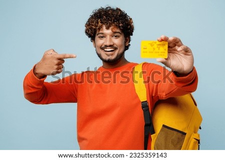 Young fun teen Indian boy student wear casual clothes backpack bag hold books hold point on credit bank card isolated on plain pastel light blue cyan background. High school university college concept