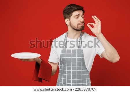 Young fun waiter male chef cook baker man wear grey apron towel hold empty plate with area making okay taste delight sign, savoring isolated on plain red background studio. Cooking food menu concept