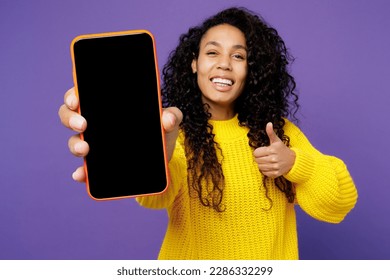 Young fun woman of African American ethnicity wear casual yellow sweater hold in hand use close up mobile cell phone with blank screen workspace area show thumb up isolated on plain purple background - Powered by Shutterstock