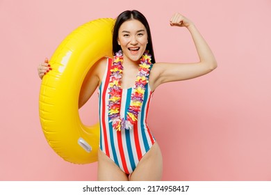 Young fun woman of Asian ethnicity in striped one-piece swimsuit hawaii lei hold inflatable ring show hand muscles isolated on plain pastel pink background. Summer vacation sea rest sun tan concept