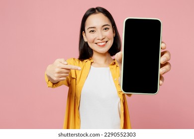 Young fun woman of Asian ethnicity wear yellow shirt white t-shirt hold use point finger on close up mobile cell phone with blank screen workspace area isolated on plain pastel light pink background