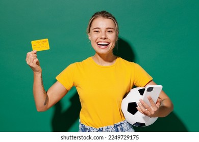 Young fun woman fan wear basic yellow t-shirt cheer up support football sport team hold in hand soccer ball mobile cell phone credit card watch tv live stream isolated on dark green background studio - Shutterstock ID 2249729175