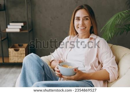 Young fun woman wear casual clothes sits in armchair eat breakfast muesli cereals with milk fruit in bowl stay at home hotel flat rest relax spend free spare time in living room indoo. Lounge concept
