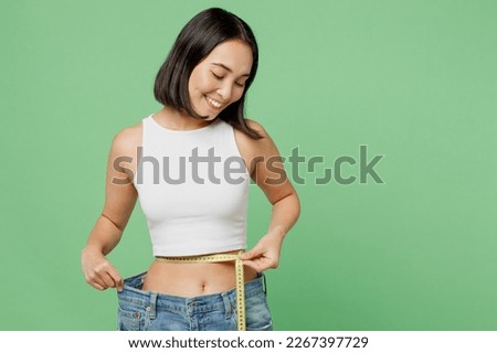 Young fun woman wear white clothes show loose pants after weightloss hold measure tape on waist isolated on plain pastel green background. Proper nutrition healthy fast food unhealthy choice concept