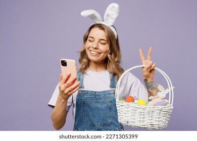 Young fun woman wearing casual clothes bunny rabbit ears hold wicker basket colorful eggs use mobile cell phone show v-sign isolated on plain purple background studio portrait. Happy Easter concept