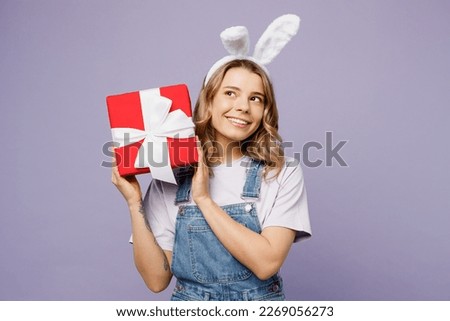 Young fun woman wearing casual clothes bunny rabbit ears hold in hand present box with gift ribbon bow isolated on plain pastel light purple background studio portrait. Lifestyle Happy Easter concept